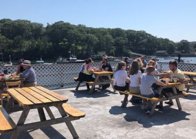People seated at picnic tables on a deck, enjoying a scenic view of the water in a relaxed outdoor setting.
